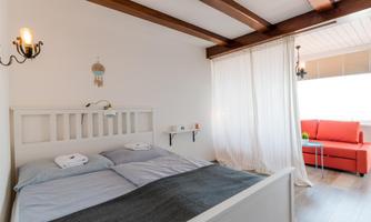 A bright bedroom with a comfortable double bed and wooden beams. In the background, there is a cozy seating area with a red couch.