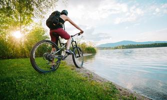 A cyclist stands by the shore of a lake and enjoys the view. The scene depicts a natural setting with trees and a calm water surface.