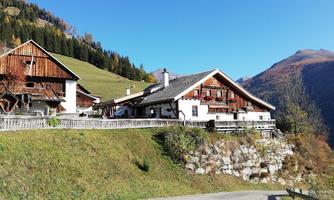 A picturesque farmhouse with traditional wooden architecture in a green, mountainous landscape. In the background, hills and mountains are visible, emphasizing the idyllic surroundings.
