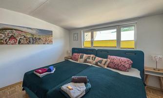 A cozy bedroom with two beds and colorful pillows. Large windows let in plenty of daylight.