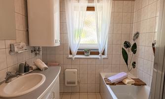 A bright bathroom with a bathtub and sink. The windows are decorated with white curtains and there are fresh plants in the room.