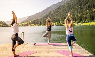 Three women are practicing yoga on a wooden terrace at the edge of a calm lake. In the background, green mountains and a clear sky can be seen.