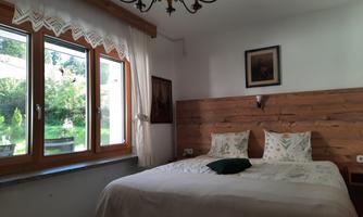 A cozy bedroom with a double bed and a window view of a garden. The walls are partially made of wood, and a decorative lamp hangs from the ceiling.