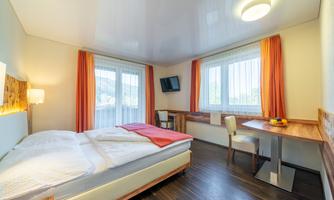 A modern hotel room with a double bed, a desk, and large windows. The walls are light, and orange curtains create a cozy atmosphere.