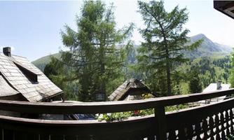 A picturesque view from a balcony of a mountain landscape. In the foreground, cozy houses and tall trees can be seen.