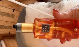 A bottle of shampoo and shower gel in orange with a golden cap. In the background, a bathtub with bubbles can be seen.