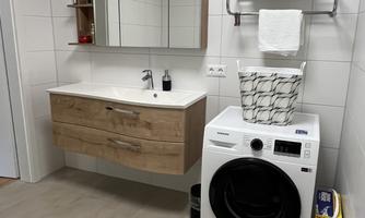 A modern bathroom with wooden furniture and a large mirror. Next to the sink is a washing machine, and towels are neatly stacked.