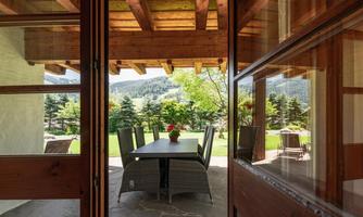 A cozy dining area with a table on a terrace, surrounded by green trees and mountains. The view through large windows creates an inviting atmosphere.