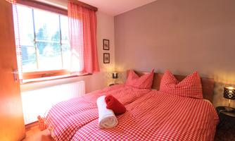 A cozy bedroom with two single beds and red plaid blankets. There is a window with plenty of daylight and decorative elements on the walls.