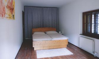 A simple bedroom with a wooden bed and two mattresses. A colorful painting hangs on the wall, and the room has bright walls and a wooden floor.