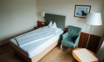 A simple hotel room with a single bed and a chair. There is a table and a picture on the wall.