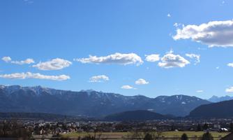 A picturesque landscape with mountains in the background and a clear blue sky. In the foreground, meadows and a small village can be seen.