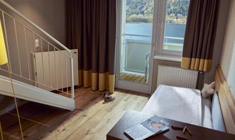 A cozy room with a large window and balcony that offers a view of the lake and mountains. The room has wooden floors and modern furniture.