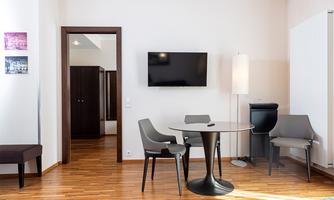 A modern room with a wooden floor and a light beige wall color. There is a table, two chairs, and a television on the wall.