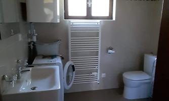 A modern bathroom with a sink, toilet, and washing machine. The room has light walls and wooden ceilings.