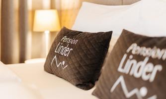 Two decorative cushions with the inscription "Pension Linder" on a bed. In the background, a soft lighting is visible.