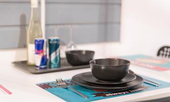 A laid table with black plates and bowls. In the background, drinks and glasses are placed on a colorful placemat.
