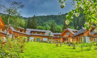 A picturesque wooden house ensemble surrounded by lush greenery and trees. The gentle landscape is overlooked by a cloudy sky.