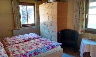 A cozy bedroom with a double bed and colorful floral bed linen. There is a wardrobe, an armchair, and a small table next to a window.