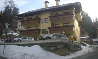 A beautiful building in alpine style with balconies. In the foreground, cars and snow can be seen.