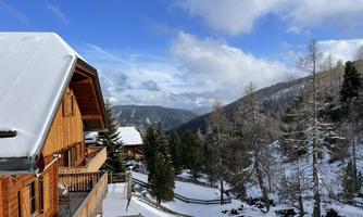 A wooden house in a snow-covered landscape with snow-covered mountains in the background. The sky is blue with some clouds, and the trees are covered with snow.