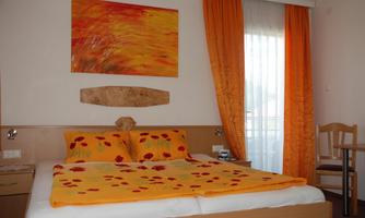 A cozy bedroom with orange curtains and a colorful wall painting. The bed is decorated with a bright yellow duvet and pillows.