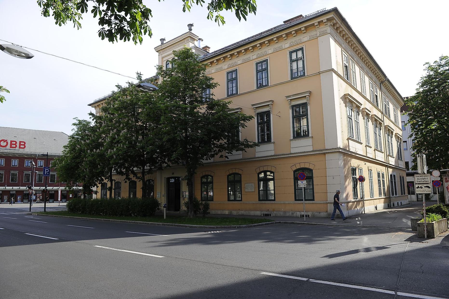 Bahnhof Mödling: Gelbes Gebäude mit Bäumen und einem Fußgänger auf der Straße