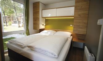 A cozy bedroom with a double bed and modern wooden details. Large windows let in plenty of daylight and offer a view of the outdoors.
