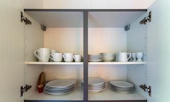 A kitchen cabinet with dishes and cutlery. On the shelves, white cups, bowls, and plates are neatly arranged.