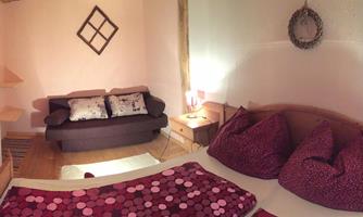 A cozy bedroom with a large bed and colorful pillows. In the background, there is a couch and soft lighting.