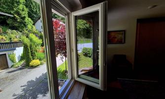 An open window with a view of a green surroundings and a garden. In the background, a few trees and an entrance can be seen.