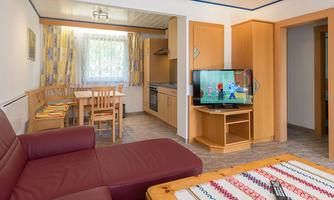 A cozy living room with a red sofa, a wooden table, and a television. The kitchen is well-equipped and the windows let in plenty of light.