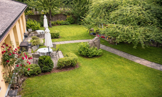 A well-maintained garden with lawn, seating areas, and two sun umbrellas. Surrounded by trees and flowers, the space offers an inviting atmosphere.
