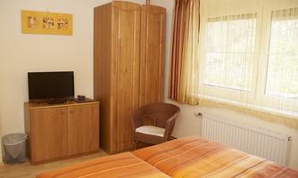 A simple bedroom with a double bed, a television, and a wooden wardrobe. The windows are draped with curtains and let in plenty of daylight.