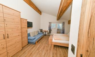 A bright room with wooden furniture and a large window. There is a double bed, a couch, and a small dining area.