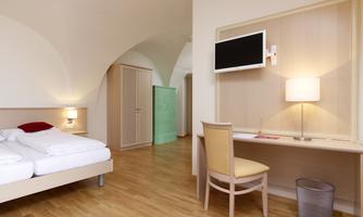 A modern hotel room with a double bed, a desk, and a television. The room features bright furniture and a warm wooden floor.