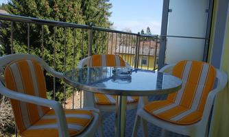 A small balcony with a round table and orange-striped chairs. In the background, there are trees and a blue sky.