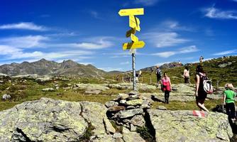A hiking route with yellow signposts on a stony landscape. In the background, there are mountains and people hiking.
