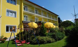 A yellow building with balconies and sunny terraces, surrounded by a well-maintained garden. A swing is in the foreground, and yellow umbrellas provide shade.