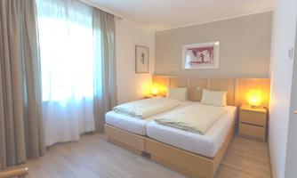 A bright and modern bedroom with two single beds. The walls are painted in soft colors, and there are large windows with curtains.