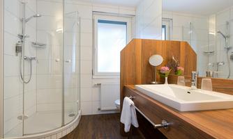 A modern bathroom with a glass shower and a stylish sink. The walls are tiled in a light color, and there are wooden details as well as some plants.