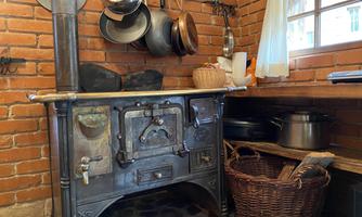 A traditional wood stove in a rustic kitchen with brick walls. Next to the stove, pots and a willow basket are visible.