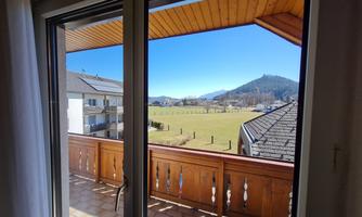 A view from a balcony over a green meadow and gentle hills. The sky is clear and blue, perfect for a relaxing day.