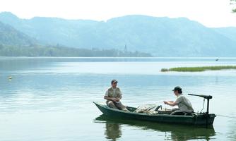 Two men are sitting in a boat on a calm lake. In the background, gentle hills and a clear sky can be seen.