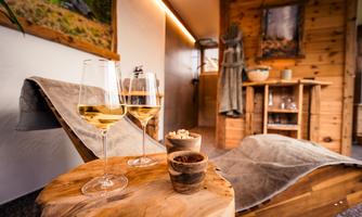 A cozy room with wooden decor, where two glasses of champagne are standing on a table. In the background, a relaxing lounge chair and a wooden console can be seen.