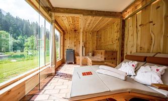 A cozy room with wooden paneling and large windows overlooking a green landscape. Two loungers and towels create a relaxing atmosphere.