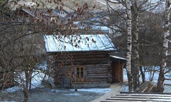 A small wooden cottage surrounded by trees and snow-covered ground. Other buildings are visible in the background.