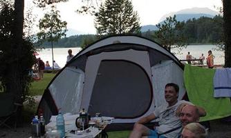 A group of people is sitting relaxed in front of a tent by the lake. Trees surround the campsite and in the background, the water is visible.