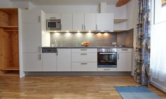 A modern kitchen with white cabinets and wooden flooring. It features a stove, a refrigerator, and a bright window front.