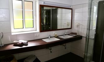 A modern bathroom with a double sink and large mirrors. The shower and a window provide brightness in the room.